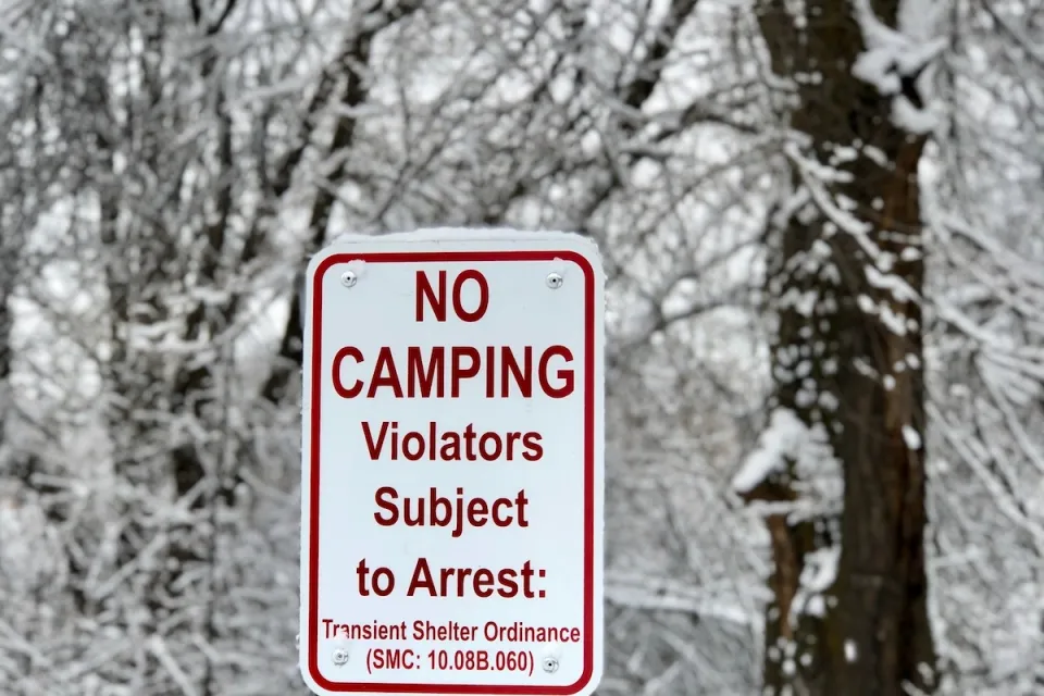 White sign with No Camping - Violators Subject to Arrest and municipal code in red text with snowy trees in background.